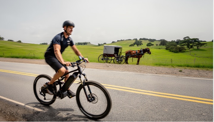 A man rides a modern electric bicycle past a horse-drawn buggy on a rural road, symbolizing progress through operations science and technology.